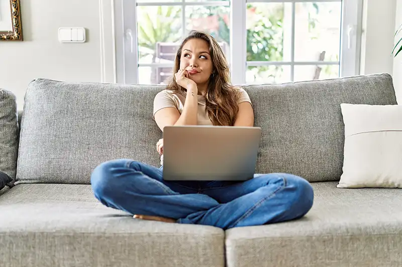 Junge Frau sitzt nachdenklich mit Laptop auf dem Sofa – sie informiert sich über Zahnersatzversicherung und vergleicht Tarife online.