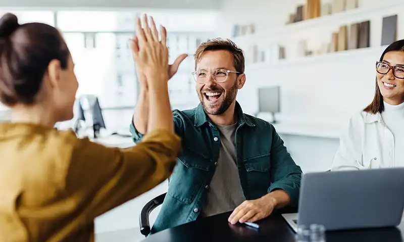 Drei berufstätige Personen in einem modernen Büro feiern gemeinsam einen Erfolg mit einem High-Five – ihr Lächeln zeigt gesunde, gepflegte Zähne. Symbolbild für Selbstbewusstsein und Ausstrahlung im Job durch gute Zahngesundheit.