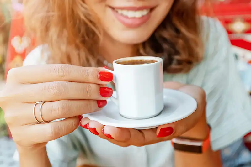 Lächelnde Frau mit weißen Zähnen hält eine kleine Tasse Kaffee mit roten lackierten Fingernägeln.